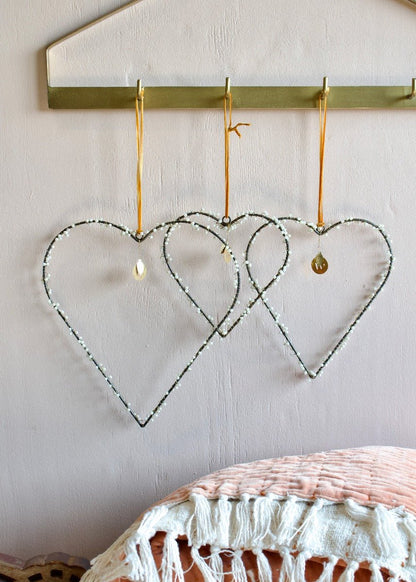 Three pearl-beaded heart ornaments hanging from gold hooks against a soft pink wall