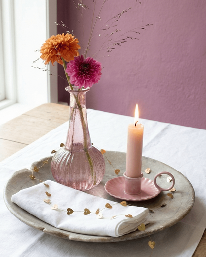 Decorative setting with a pink vase, candle, and flowers on a white tablecloth.