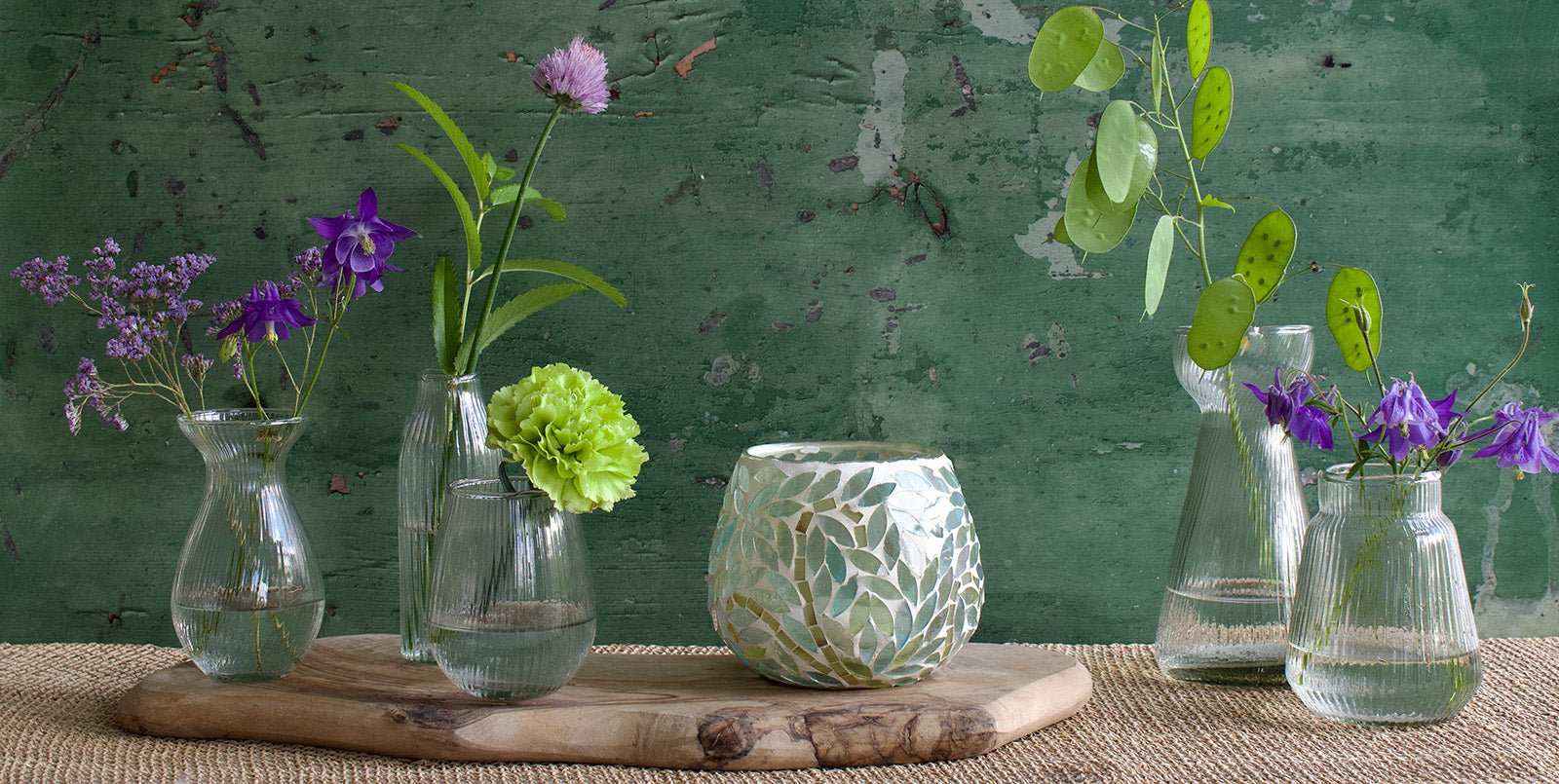 A collection of ribbed glass vases in various shapes filled with fresh flowers, arranged on a wooden tray alongside a round mosaic tealight holder. The setting features a rustic green wall in the background and a woven placemat underneath.