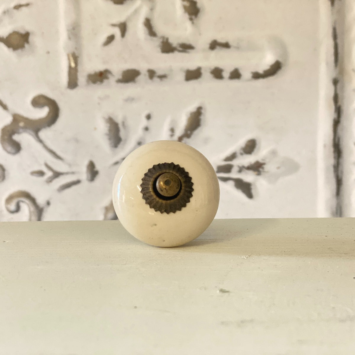 A white ceramic furniture knob with a metallic design inserted in the center, placed against a white background with decorative patterns.
