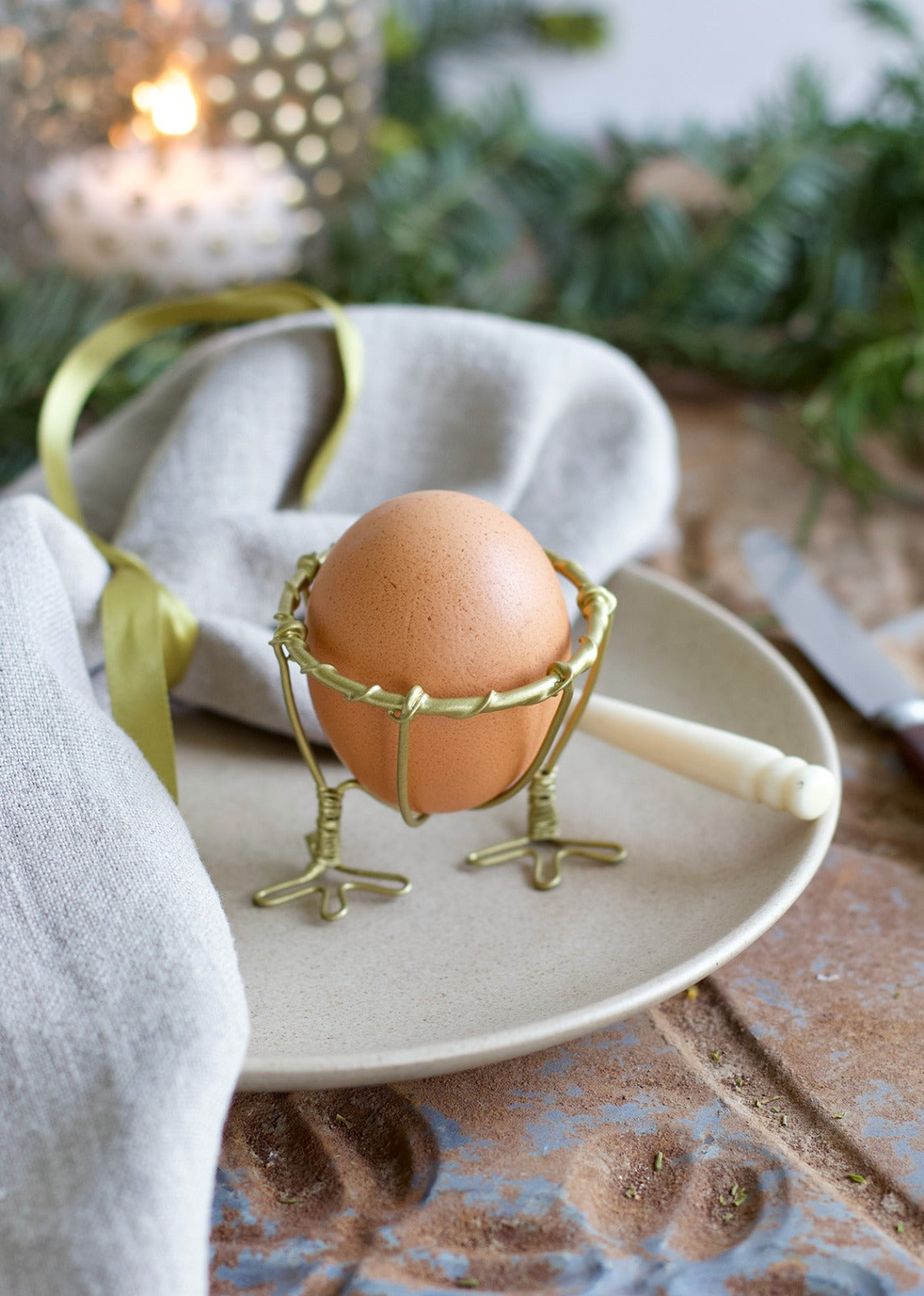 Egg in a decorative metal holder on a plate with a candle and greenery in the background
