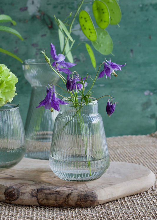 Small ribbed glass vase with a soft round shape, filled with water and delicate purple flowers, placed on a wooden board.