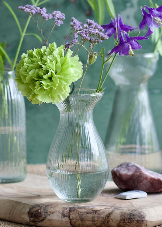 A pear-shaped ribbed glass vase filled with water and delicate spring flowers, including a vivid green carnation and purple statice. The vase sits on a wooden plank with natural stones, set against a rustic green background for a soft and organic look.