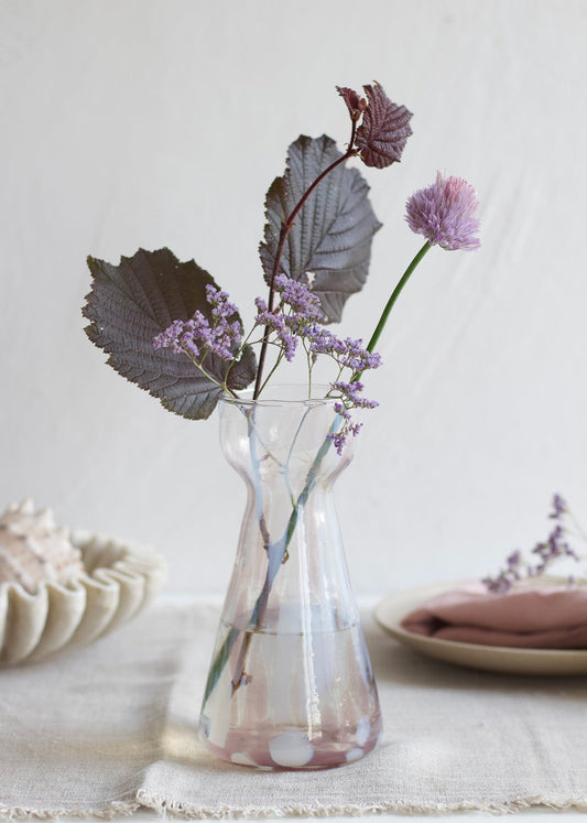 Handmade pink glass vase with soft white spots and hourglass shape, styled with purple flowers and dark foliage on a linen table.
