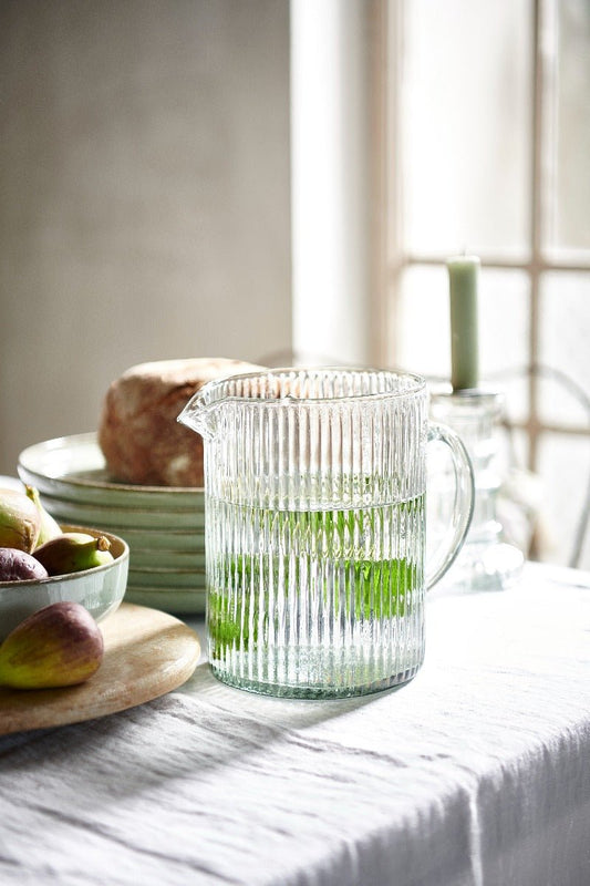 Handblown ribbed glass pitcher in champagne colour, filled with water and mint, placed on a softly lit table with figs, bread, and ceramic plates in the background.