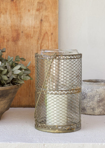 Decorative metal lantern with a candle inside, placed on a surface with a wooden board and plant in the background.