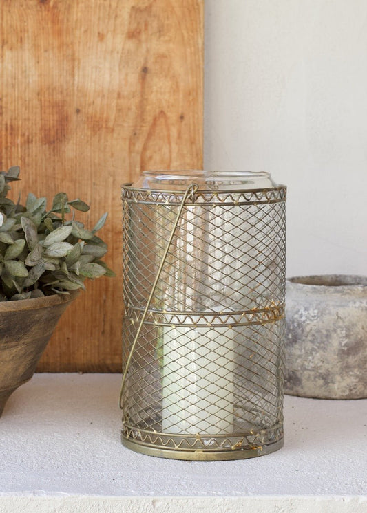 Decorative metal lantern with a candle inside, placed on a surface with a wooden board and plant in the background.
