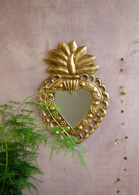 Heart-shaped brass wall mirror with flame crown and dotted floral details, hanging on a pink textured wall.