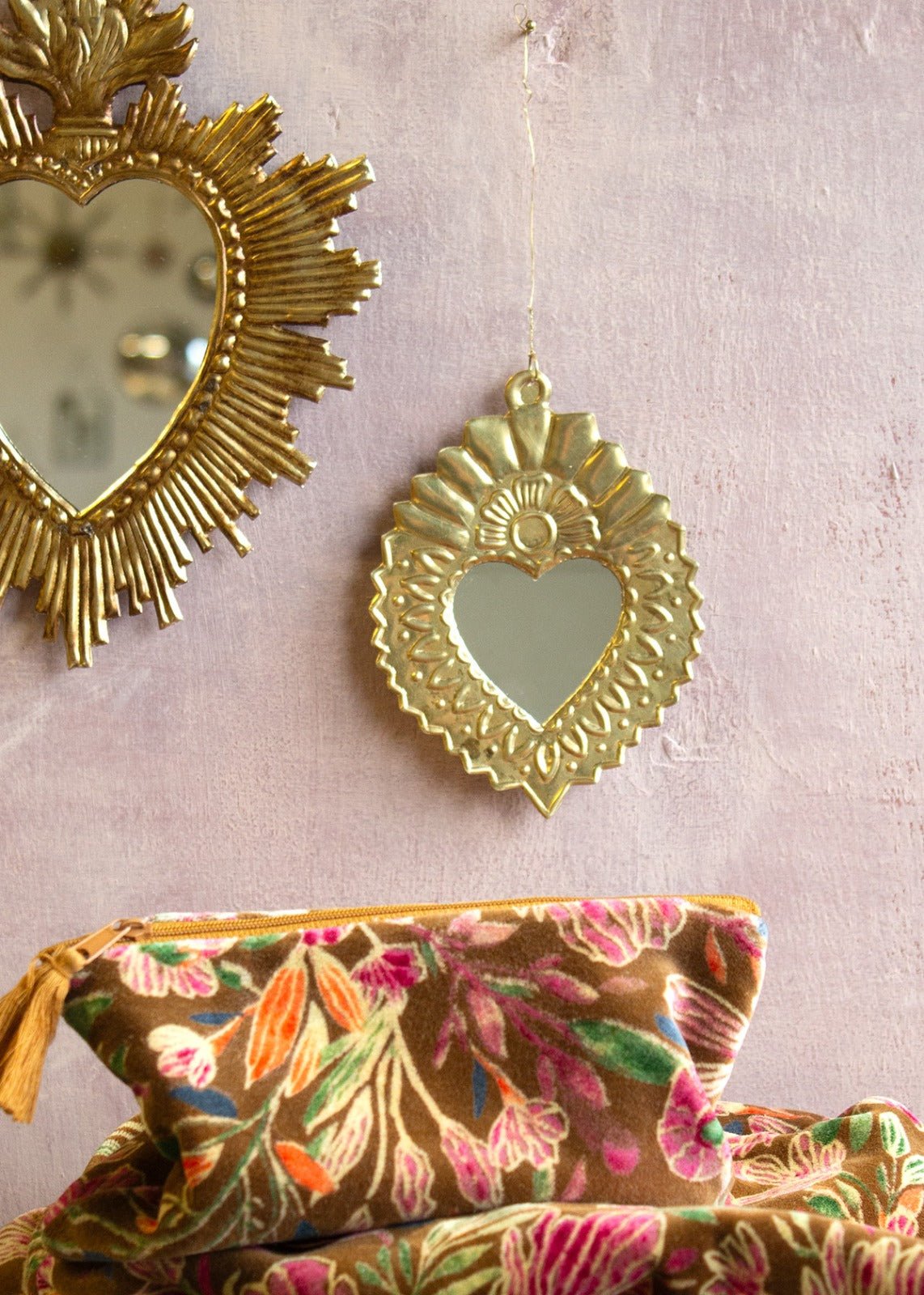 Gold-toned heart-shaped mirror with fan-shaped detailing at the top, hanging on a pink wall with a floral pouch in the foreground.
