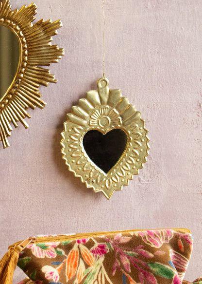 Small heart-shaped ex-voto mirror in shiny brass with embossed floral and sunburst details, hanging on a soft pink wall.