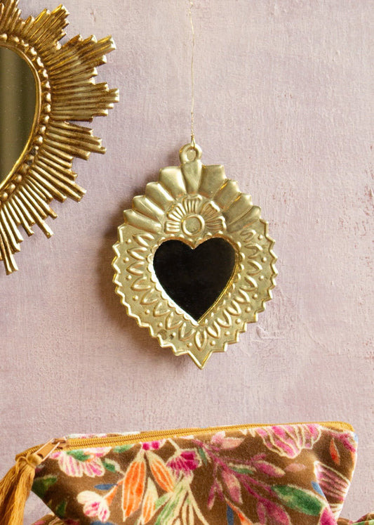 Small heart-shaped ex-voto mirror in shiny brass with embossed floral and sunburst details, hanging on a soft pink wall.