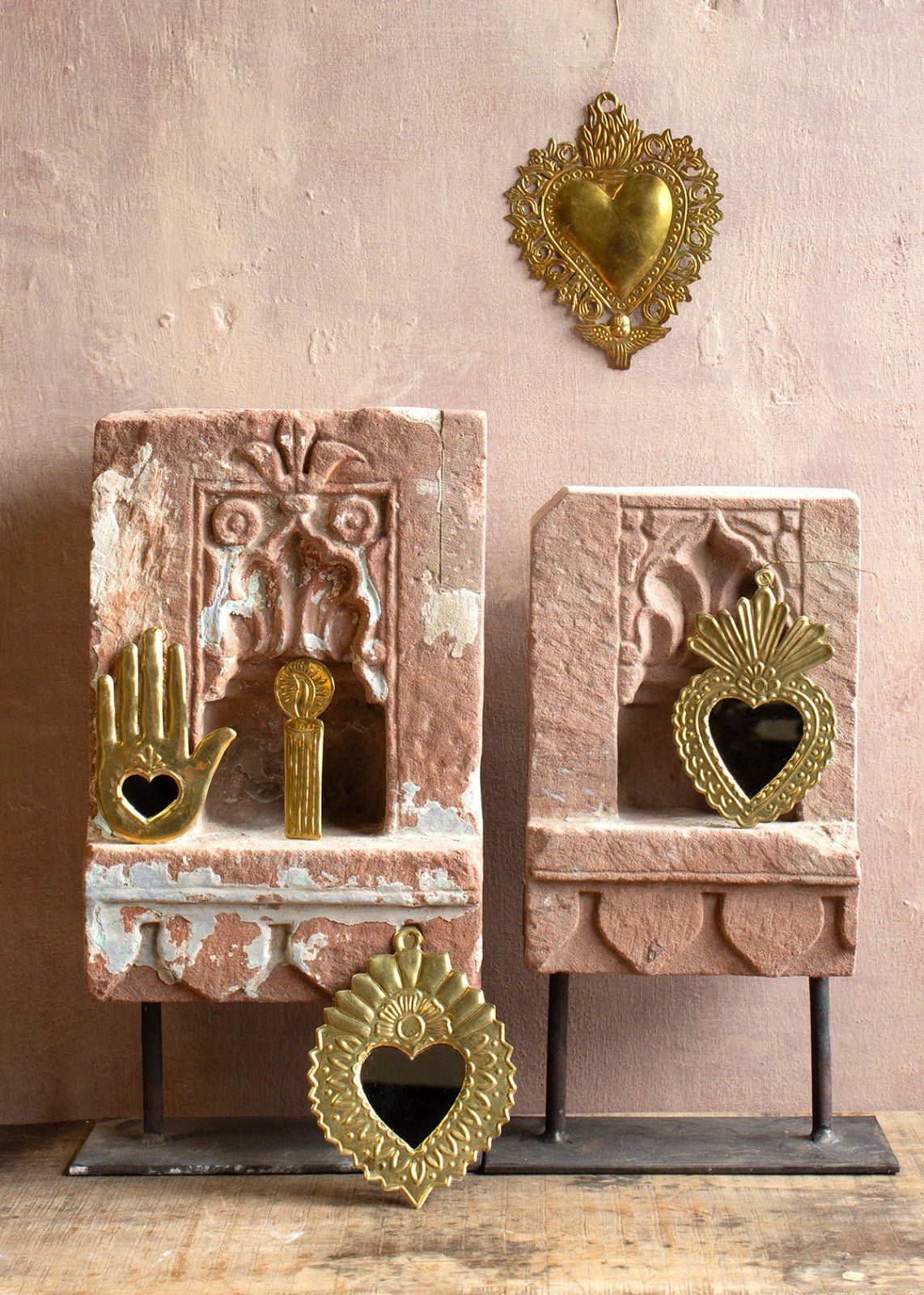 Decorative handmade metal wall ornaments, including heart-shaped mirrors with gold-colored frames, displayed in and on pink stone niches against a dusty rose wall.