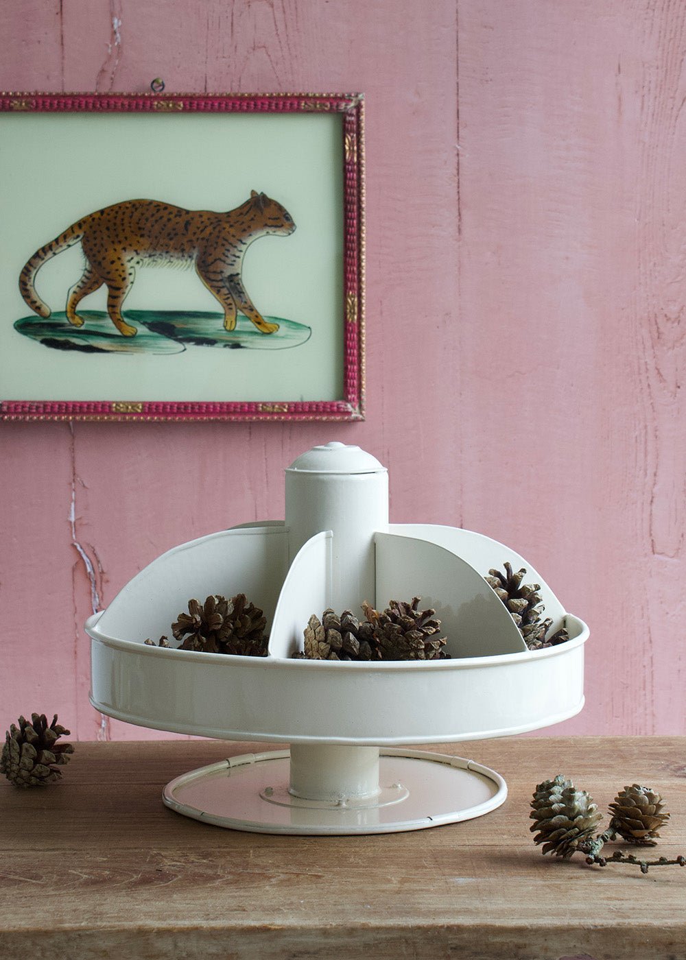 White vintage-style rotating enamel organizer with compartments, filled with pinecones, displayed on a wooden table in front of a pink wall.