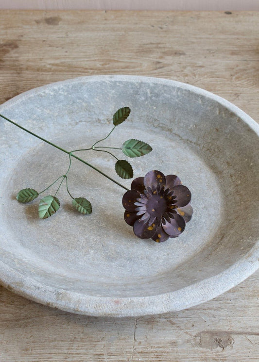 Dark brown handmade metal flower with layered petals and green leaves, displayed on a rustic stone plate.