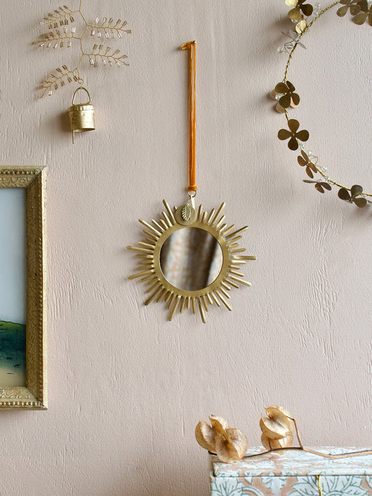 Small round brass mirror with radiant sunburst rays, hanging from an amber velvet ribbon on a textured blush pink wall, surrounded by decorative golden wall ornaments and a framed painting.