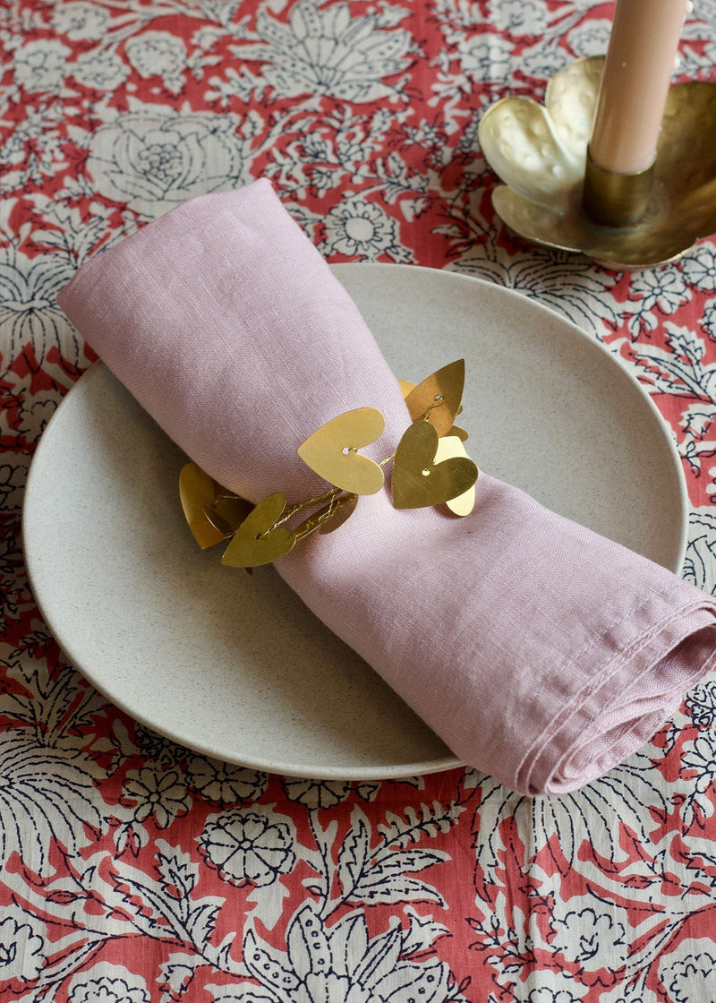 Pink napkin rolled up with gold leaf decorations on a white plate, placed on a floral tablecloth.