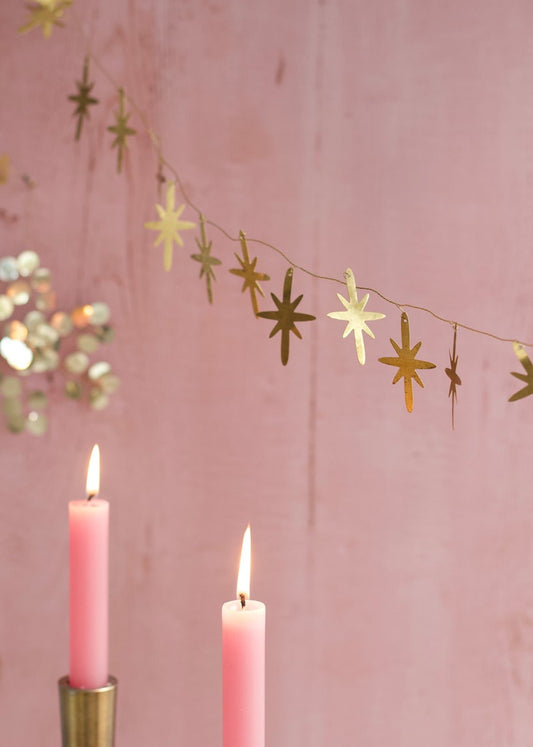 Golden metal star garland hanging against a soft pink wall, with two lit pink candles in brass holders in the foreground.