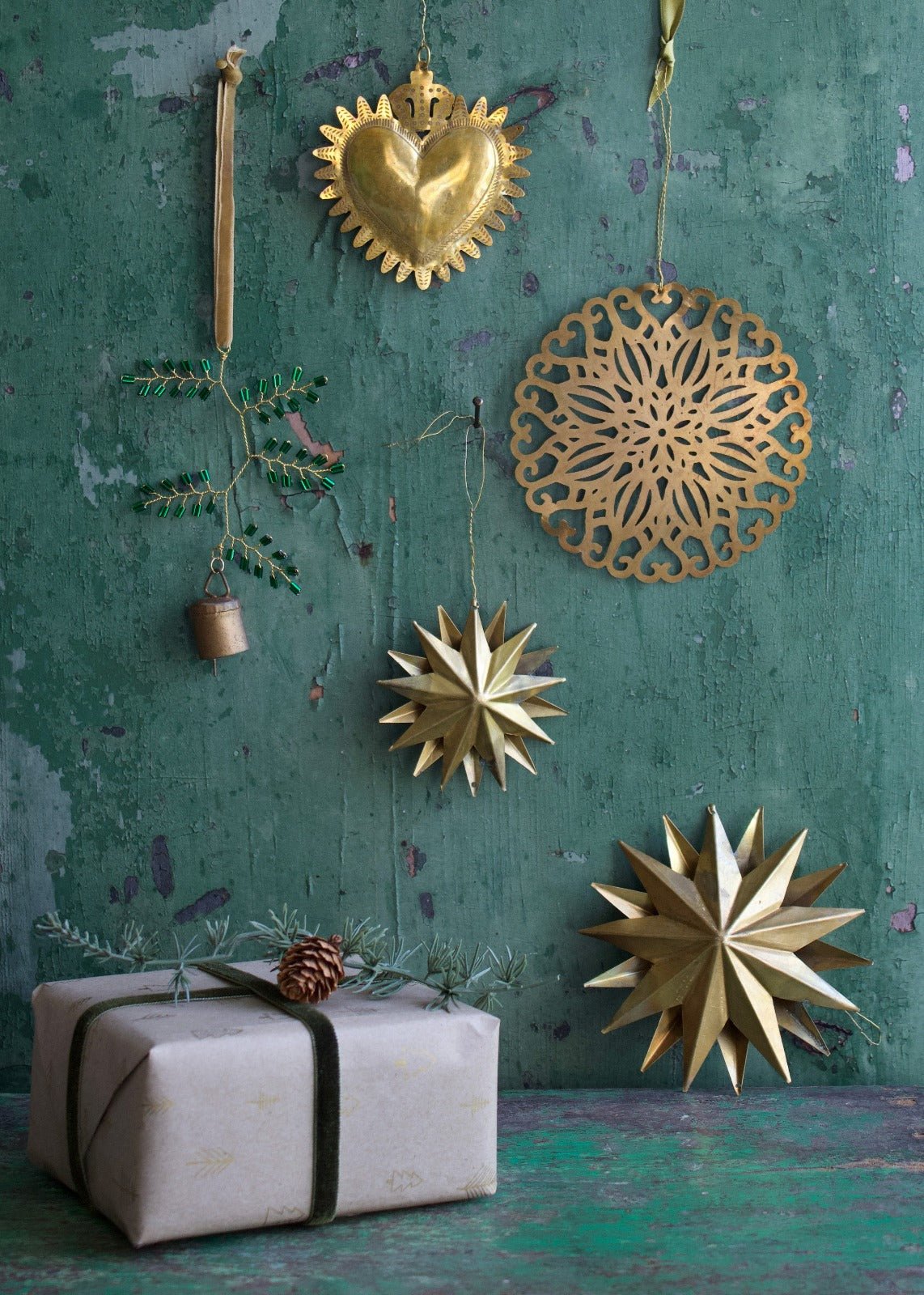 Festive arrangement of handmade brass Christmas ornaments, including a sacred heart, a round cut-out motif, golden stars, and a decorative green twig with bell, displayed against a weathered green wooden background with a pine-decorated gift.
