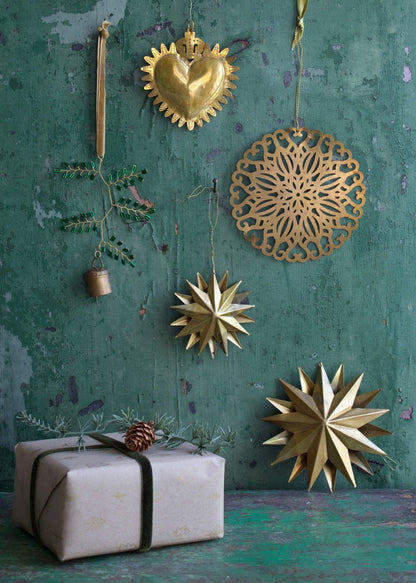 Festive arrangement of handmade brass Christmas ornaments, including a sacred heart, a round cut-out motif, golden stars, and a decorative green twig with bell, displayed against a weathered green wooden background with a pine-decorated gift.