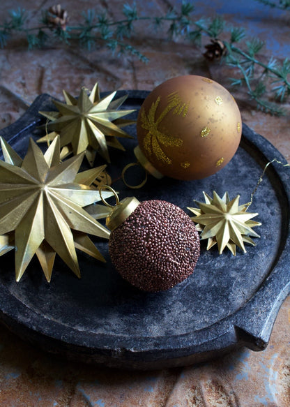 Festive brass star ornaments and decorative baubles displayed on a rustic black tray with evergreen sprigs in the background.