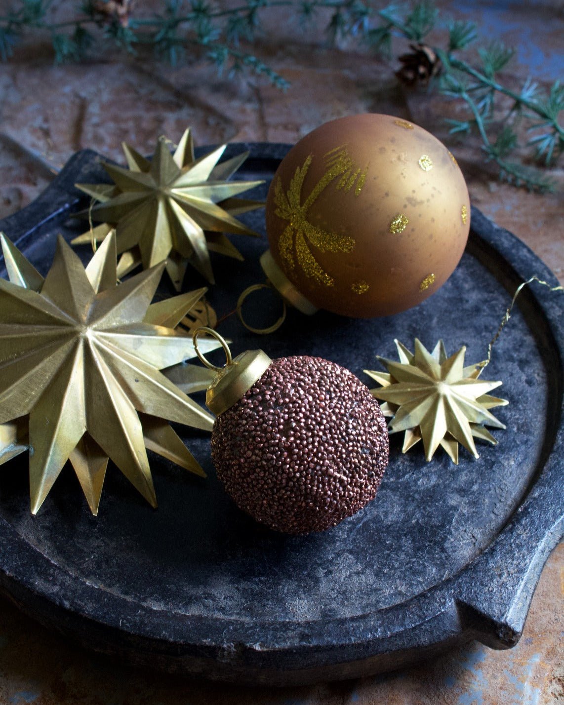 Festive brass star ornaments and decorative baubles displayed on a rustic black tray with evergreen sprigs in the background.

