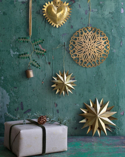 Festive arrangement of handmade brass Christmas ornaments, including a sacred heart, a round cut-out motif, golden stars, and a decorative green twig with bell, displayed against a weathered green wooden background with a pine-decorated gift.