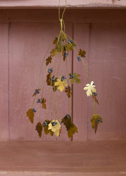Hanging brass bunch with oak-shaped leaves and grey beads, displayed against a soft pink wooden background.