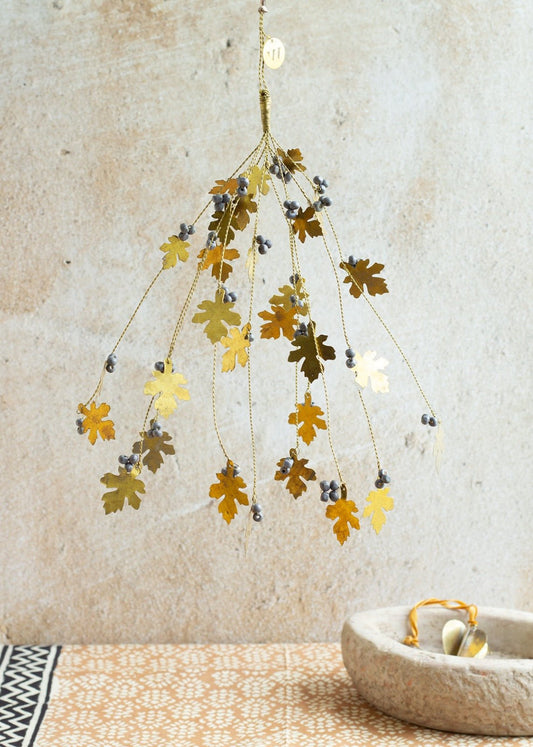 Decorative brass bunch with oak leaf shapes and grey beads, hanging against a textured beige wall above a patterned surface with a stone bowl.