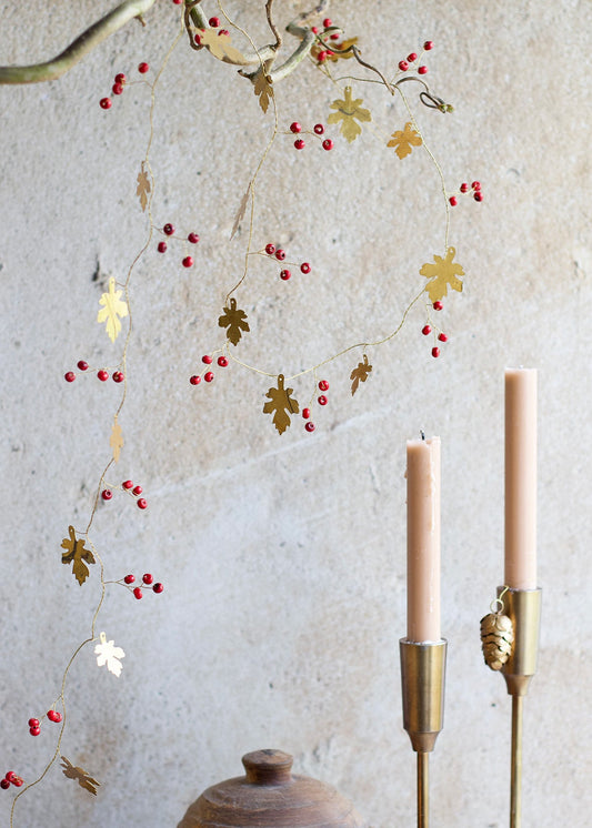 Festive garland with golden brass oak leaves and small red berries, hanging from a branch above two brass candleholders with peach-colored candles, set against a textured beige wall.