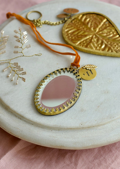 Oval ex voto mirror ornament with golden frame and orange velvet ribbon, styled on a marble plate with pink fabric background.