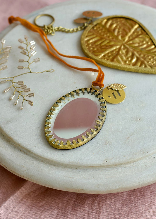 Oval ex voto mirror ornament with golden frame and orange velvet ribbon, styled on a marble plate with pink fabric background.