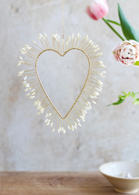 Delicate heart-shaped ornament made of gold wire and ivory-colored buds, hanging in front of a soft background with tulips.