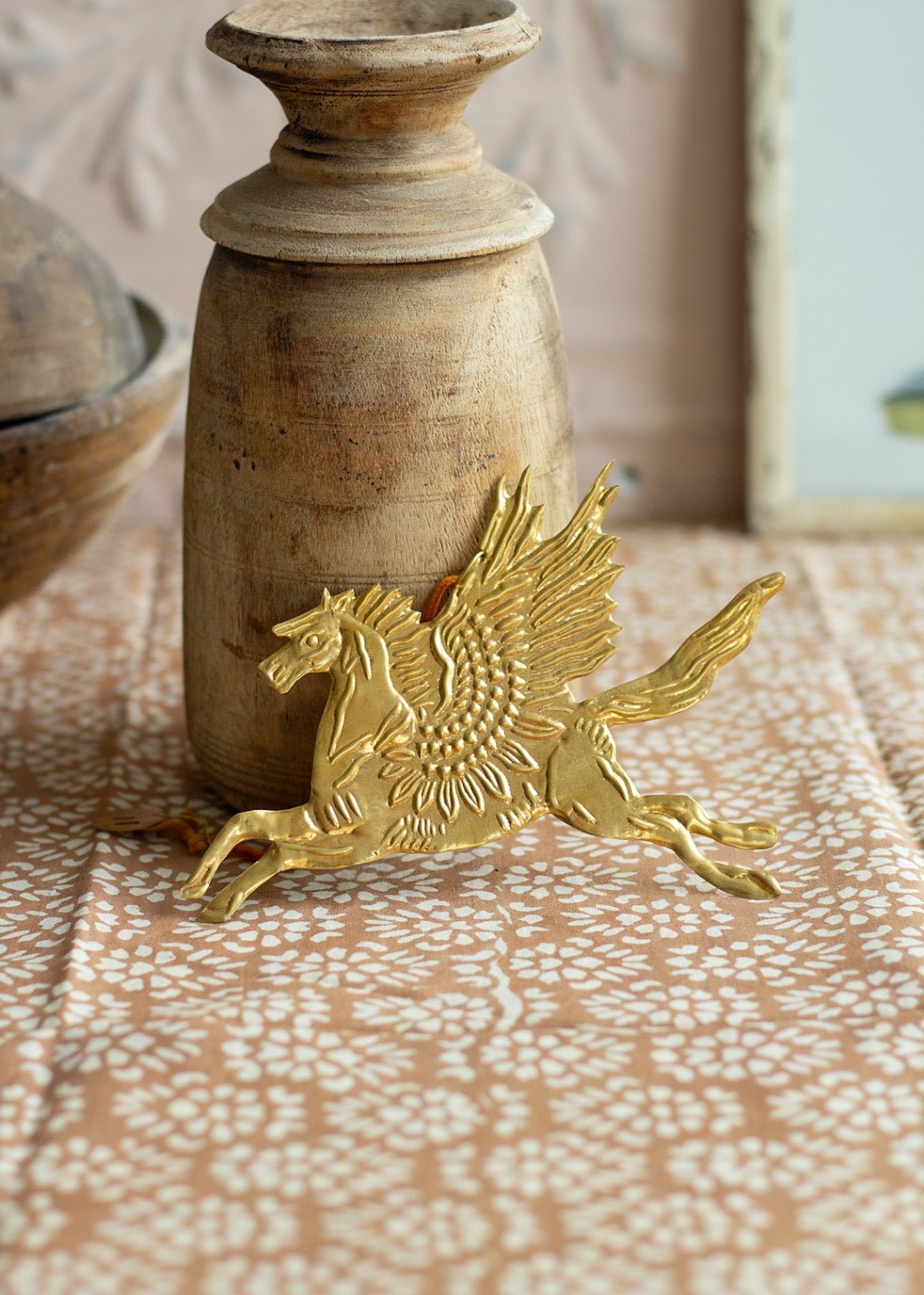 Gold-colored brass ornament of a winged horse (Pegasus) lying on a patterned textile next to a rustic wooden candle holder