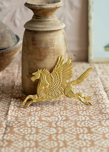 Gold-colored brass ornament of a winged horse (Pegasus) lying on a patterned textile next to a rustic wooden candle holder