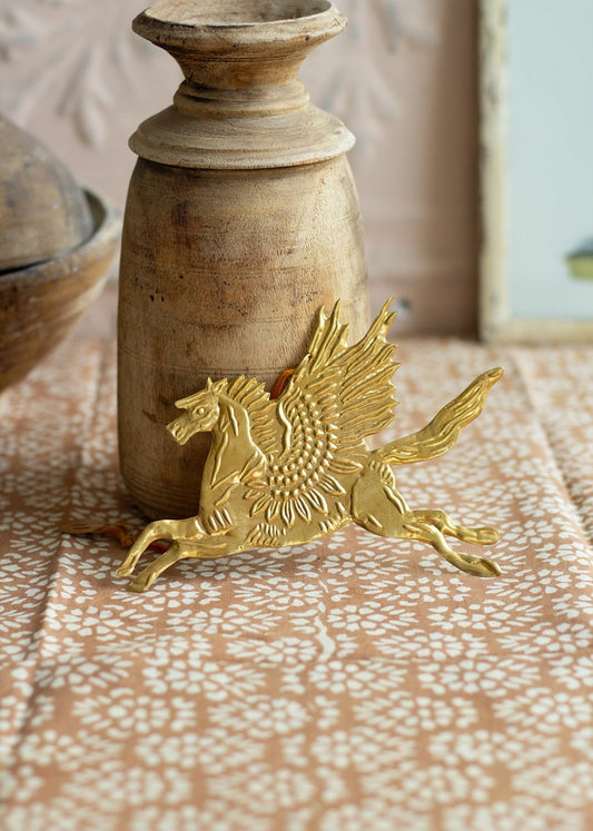 Gold-colored brass ornament of a winged horse (Pegasus) lying on a patterned textile next to a rustic wooden candle holder