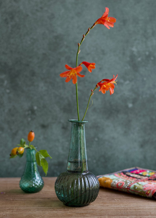 Two green glass vases with orange flowers on a wooden surface against a gray background