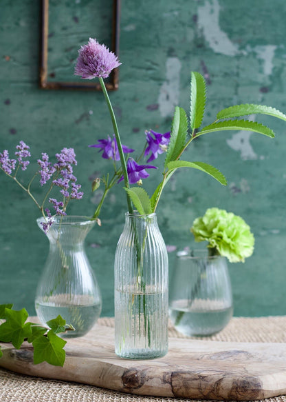 Three small glass vases with flowers on a wooden surface against a green textured wall.