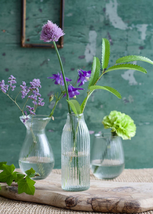 Three small glass vases with flowers on a wooden surface against a green textured wall.
