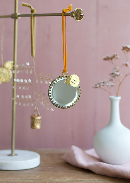 Round ex voto mirror ornament with golden frame and orange velvet ribbon, styled on a brass ornament stand with a pink background.
