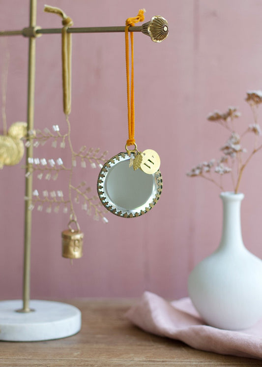 Round ex voto mirror ornament with golden frame and orange velvet ribbon, styled on a brass ornament stand with a pink background.