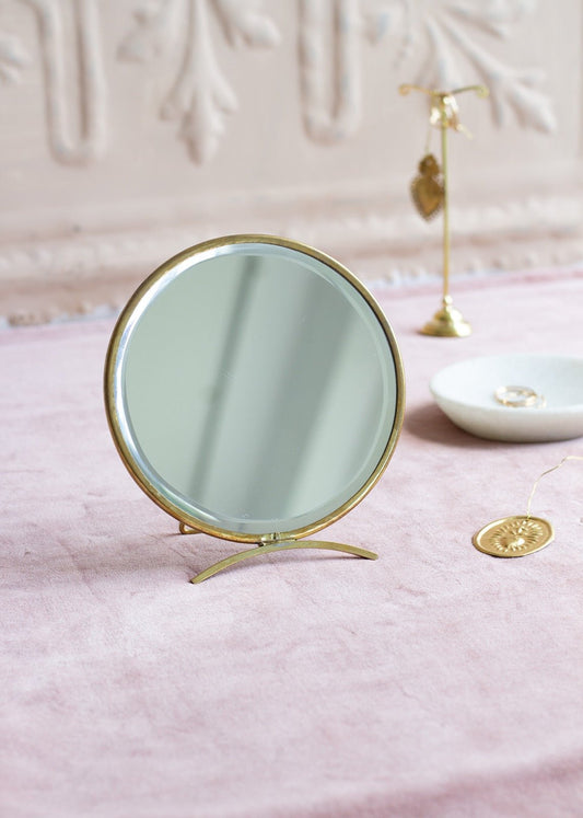 Round table mirror with brass frame and curved stand, placed on a soft pink fabric with golden jewellery and decor in the background.