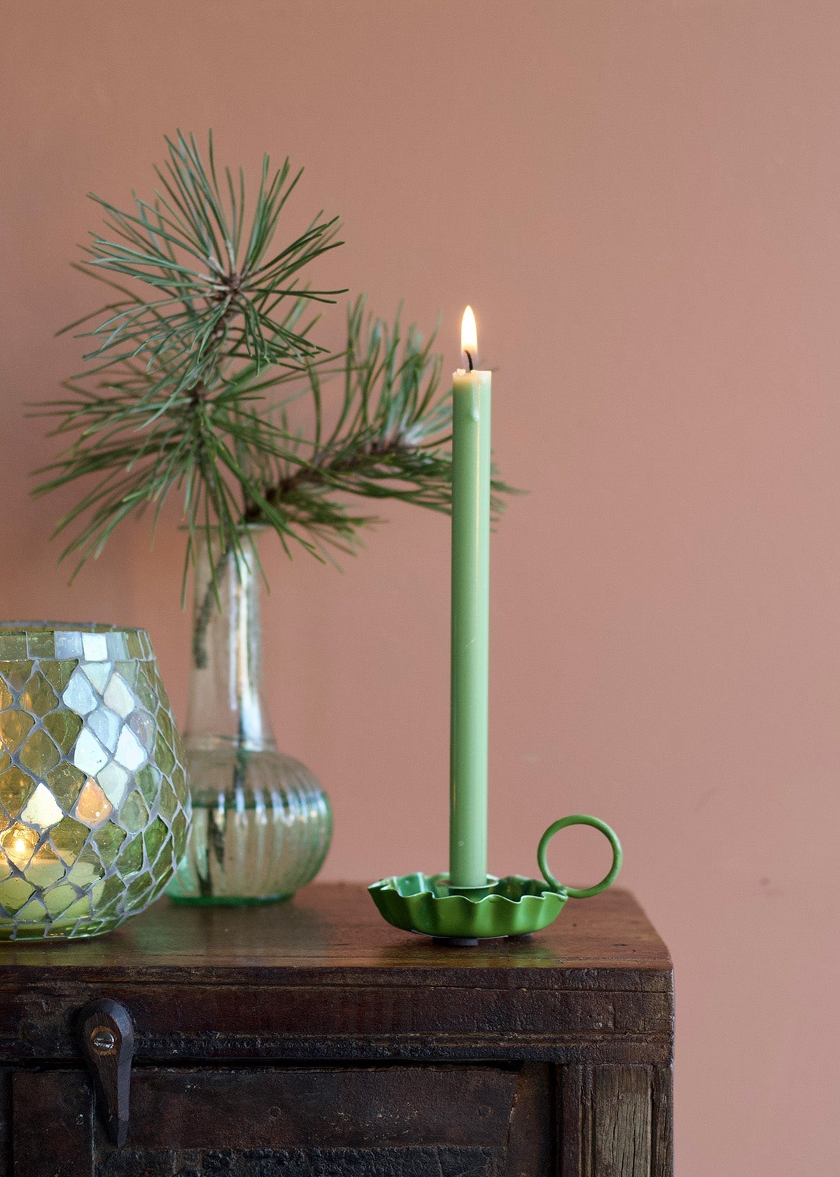 Green candle in a decorative holder with a plant and glass vase on a wooden surface against a pink wall.