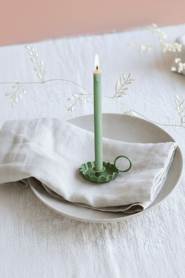 Green candle in a decorative holder on a white plate with a white napkin, set against a light pink background.