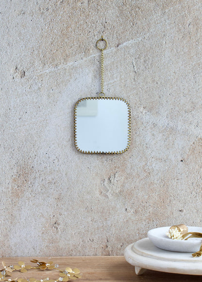 Small square mirror with rounded corners, brass edge and decorative chain for hanging, styled on a light stone wall with delicate accessories.