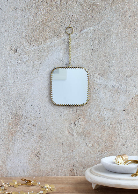 Small square mirror with rounded corners, brass edge and decorative chain for hanging, styled on a light stone wall with delicate accessories.