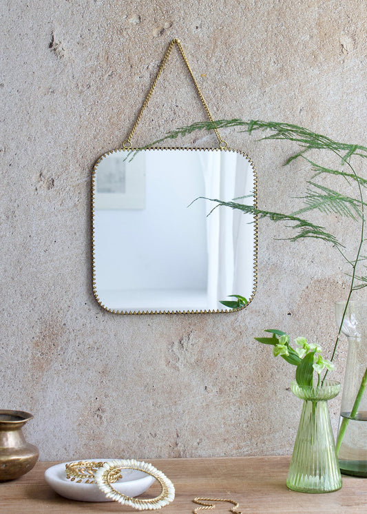 Square mirror with rounded corners and a brass scalloped edge, hanging from a brass chain on a rustic wall, styled with soft green flowers and vintage decor.