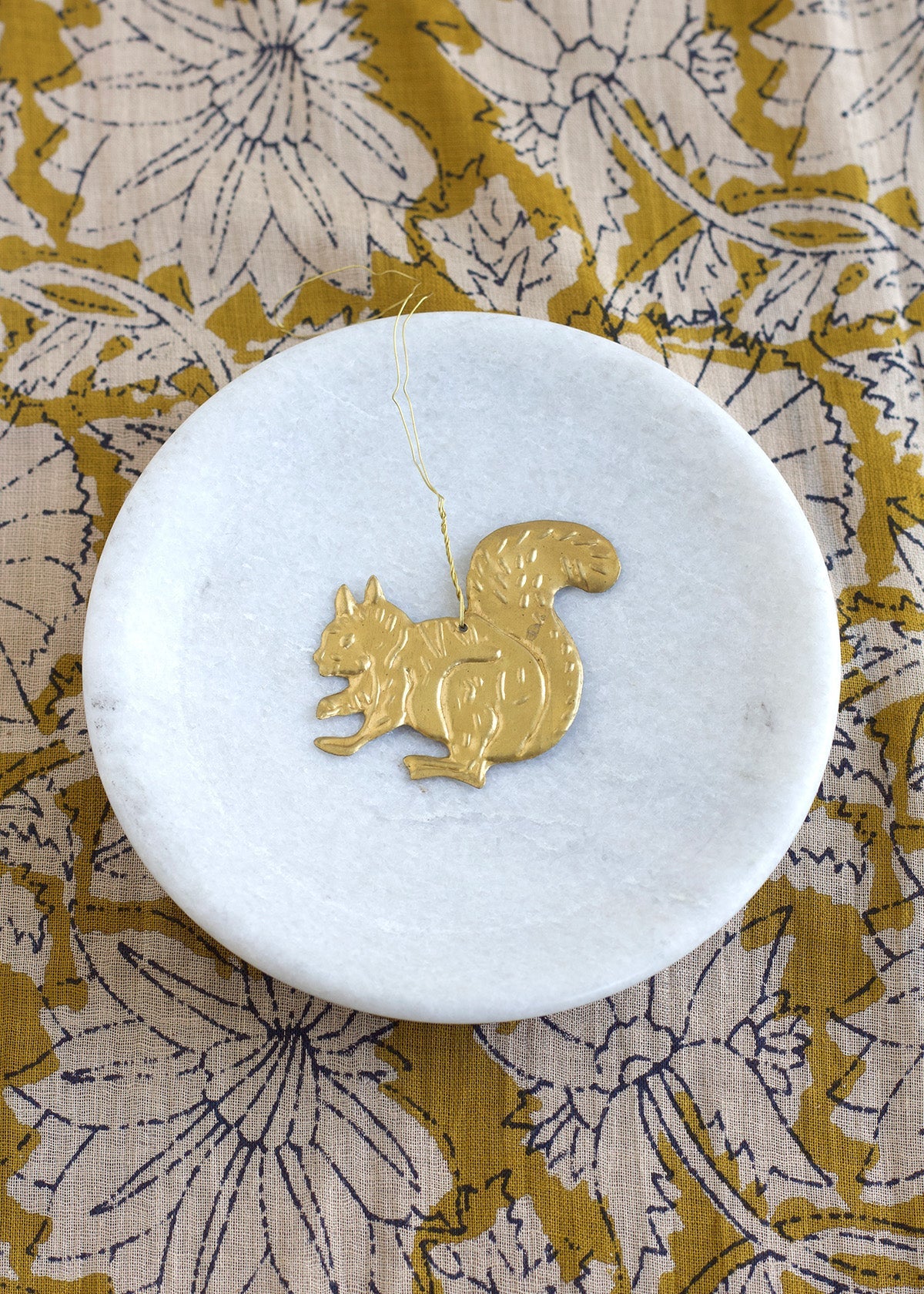 Gold squirrel pendant on a white marble coaster with a floral patterned fabric background