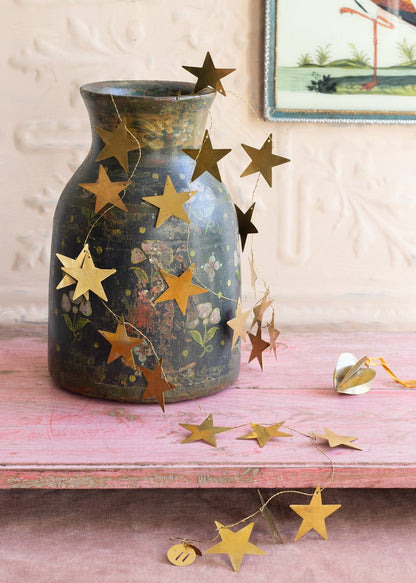 Golden brass star garland draped over a vintage painted vase with floral motifs, resting on a pink wooden surface with a soft pastel backdrop and a small heart ornament nearby.