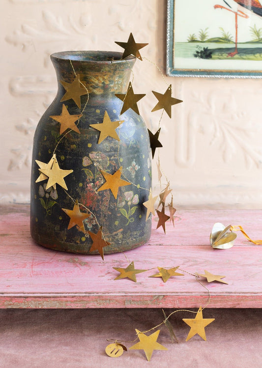 Golden brass star garland draped over a vintage painted vase with floral motifs, resting on a pink wooden surface with a soft pastel backdrop and a small heart ornament nearby.