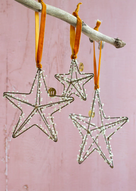 Three handmade star-shaped ornaments made of wire and tiny white beads, hanging from ochre velvet ribbons on a driftwood branch against a soft pink background.